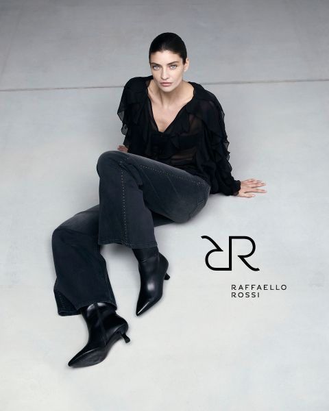 Une femme aux cheveux foncés est assise sur un sol gris clair et porte un chemisier noir à volants, un jean foncé et des bottes noires pointues. À côté d'elle, on peut voir le logo de Raffaello Rossi.