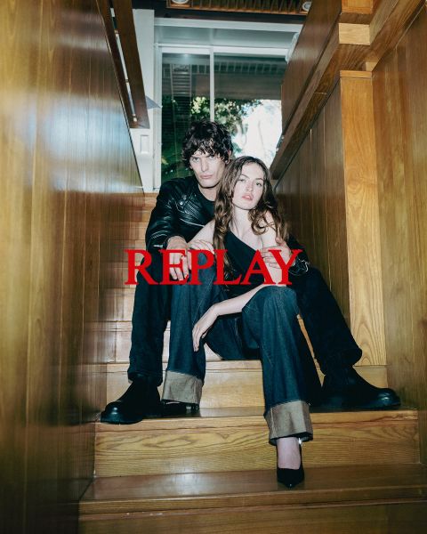 Un homme et une femme, vêtus de vêtements sombres et élégants, sont assis sur un escalier en bois dans un intérieur. Le mot "REPLAY" est affiché bien en évidence au centre de l'image en lettres capitales rouges.