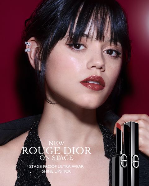 Une femme portant un rouge à lèvres rouge foncé brillant pose devant un fond rouge profond, vêtue d'un haut dos nu noir scintillant et de manchettes d'oreille. Elle tient un tube de Rouge Dior Shine Lipstick. Le texte fait la promotion du nouveau rouge à lèvres ultra-tenue, résistant à la scène.