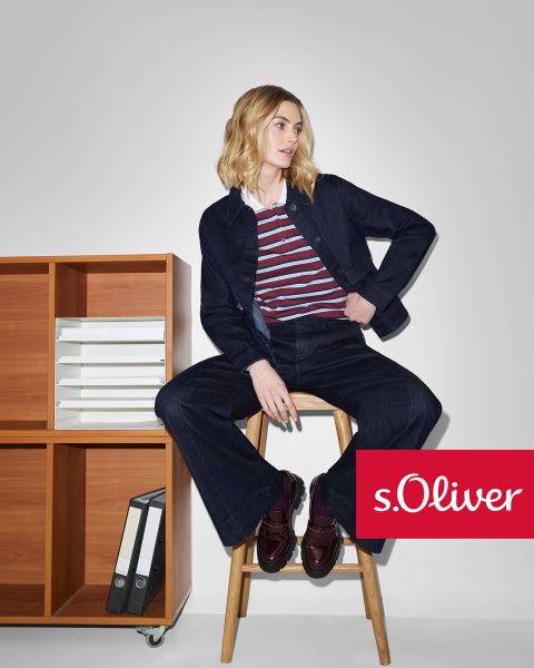 Une femme portant une veste et un pantalon en jean foncé, avec un haut rayé, est assise sur un tabouret en bois à côté d'une étagère en bois contenant des dossiers. Le logo s.Oliver apparaît dans le coin inférieur droit.