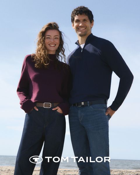 Une femme souriante portant un pull marron et un homme portant un pull bleu marine se tiennent côte à côte sur une plage. Ils portent tous les deux des jeans. Le ciel est clair et bleu. Le logo "Tom Tailor" apparaît en bas.