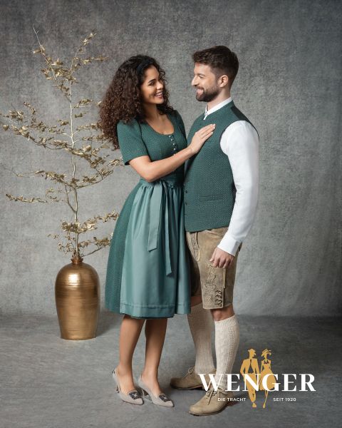Une femme souriante en dirndl vert se tient à côté d'un homme en costume traditionnel bavarois, comprenant une culotte de peau et un gilet. Ils se font face devant un fond de studio avec un vase doré et une branche décorative. Logo "Wenger" en bas.