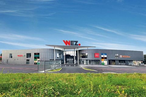 Un centre commercial moderne avec un grand panneau "WEZ" sur le toit, une façade grise, diverses enseignes de magasins et un ciel bleu clair au-dessus.