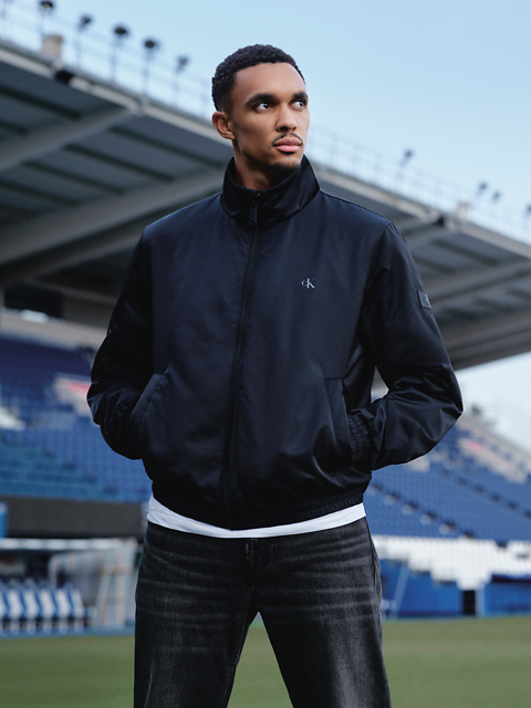 Un homme en veste noire et jean se tient dans un stade, les mains dans les poches, et regarde sur le côté. On aperçoit des sièges de stade et un grand bâtiment en arrière-plan.