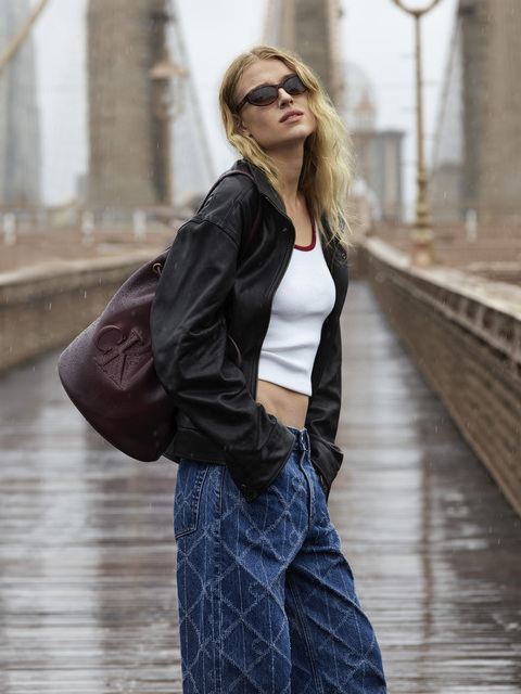 Une femme portant des lunettes de soleil, une veste noire, un crop-top blanc et un jean bleu ample se tient sur un pont pluvieux et porte un grand sac marron sur l'épaule. Le pont et les lampes sont flous à l'arrière-plan.