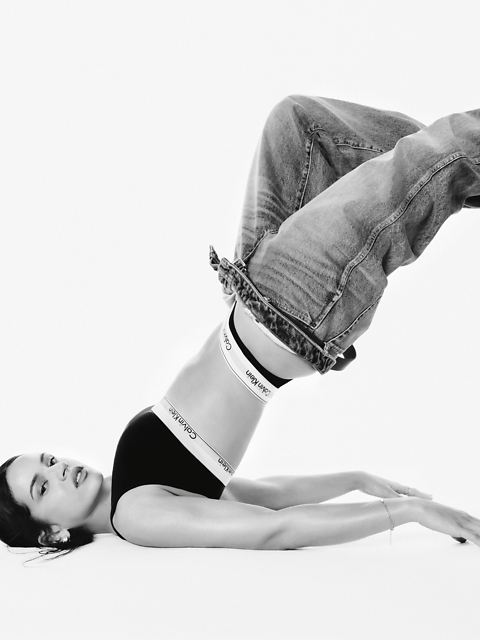 Photo en noir et blanc d'une femme portant un soutien-gorge de sport Calvin Klein et un jean, allongée sur le dos, les jambes pliées au-dessus de la tête, les bras tendus sur le sol, le regard fixé sur l'appareil photo.