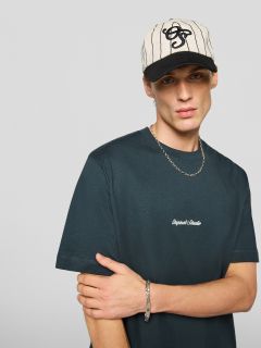 Un jeune homme portant un t-shirt noir avec l'inscription "Original Studio", un collier argenté, un bracelet argenté et une casquette de baseball rayée crème et noire se tient devant un fond gris clair uni.