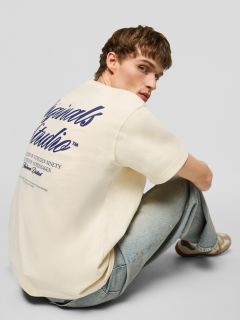 Un jeune homme aux cheveux bruns courts, portant un t-shirt crème avec un imprimé bleu marine dans le dos, un jean bleu clair et des baskets beiges, est assis par terre et regarde par-dessus son épaule en direction de la caméra.