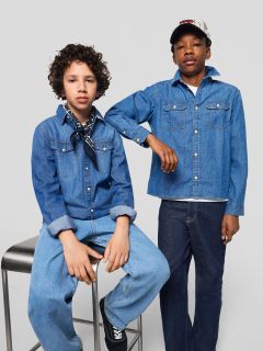 Deux garçons en jeans et chemises en jean posent ensemble. L'un est assis sur un tabouret, les mains sur les genoux et une écharpe autour du cou, tandis que l'autre, le bras posé sur le tabouret, porte un bonnet.