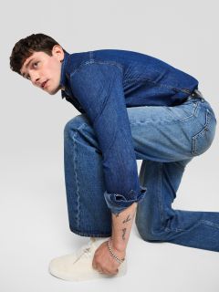 Un jeune homme en veste en jean et jean est agenouillé et regarde sur le côté. Il porte des baskets blanches et a un tatouage visible sur son avant-bras. L'arrière-plan est uni, gris clair.