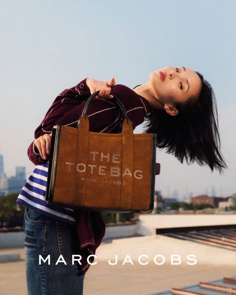 Une femme pose en extérieur avec un sac marron Marc Jacobs à la main, se penche en arrière la tête inclinée, portant une veste marron et un jean bleu. Les mots "THE TOTE BAG MARC JACOBS" et "MARC JACOBS" apparaissent sur l'image.