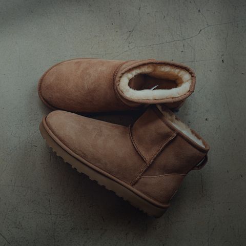 Une paire de bottes d'hiver en daim marron clair avec une épaisse doublure en polaire claire, posées sur un sol en béton gris légèrement fissuré.