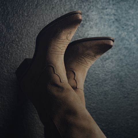 Gros plan d'une paire de bottes de cowboy marron en daim avec des coutures décoratives sur un fond sombre et texturé. Les bottes sont orientées vers le haut, mettant en valeur leur design et leur matière.