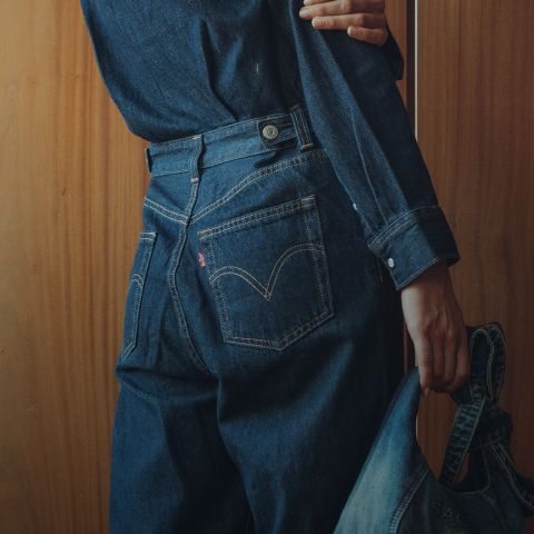 Une personne portant une chemise en jean foncé et un jean se tient devant un mur en bois et tient une veste en jean dans une main, tout en tournant le dos à la caméra.