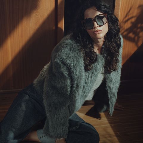 Une femme aux cheveux ondulés et foncés, portant des lunettes de soleil surdimensionnées, une veste de fourrure grise et un jean foncé, est assise sur un plancher en bois près d'un mur en bois, dans un éclairage dramatique.