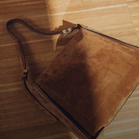 Un sac bandoulière en daim marron avec une large bandoulière est posé sur un plancher en bois ensoleillé, où la lumière et l'ombre créent un aspect chaleureux et texturé.