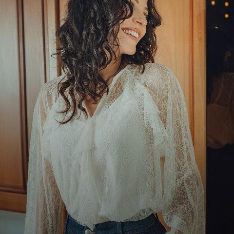 Une femme aux cheveux ondulés et foncés, portant un chemisier en dentelle blanche et un jean bleu, se tient souriante dans une pièce aux boiseries chaudes et éclairées, et regarde sur le côté.