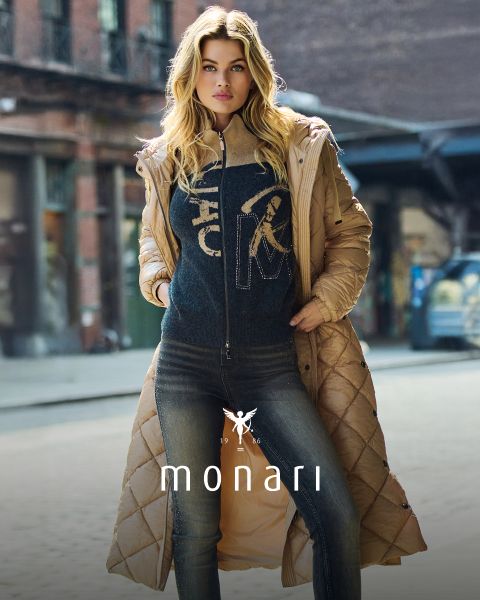 Une femme aux longs cheveux blonds porte un manteau matelassé marron clair sur un pull foncé et un jean, et se tient avec assurance dans une rue de la ville. Le mot "monari" est affiché en bas de l'image.