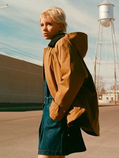 Une personne aux cheveux blonds courts se tient dans une rue et porte une veste marron sur une robe bleue. À l'arrière-plan, par ciel clair, on aperçoit un château d'eau et des lignes électriques.