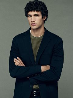 Un homme aux cheveux bouclés et foncés, portant un blazer noir sur un pull en maille côtelée verte, se tient les bras croisés devant un fond gris uni et regarde droit devant lui avec une expression neutre.