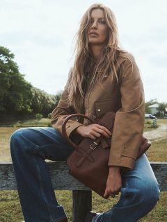 Une femme aux longs cheveux blonds est assise à l'extérieur sur une clôture en bois, portant une veste marron clair, un jean bleu et tenant un sac à main marron sur ses genoux. À l'arrière-plan, on peut voir des arbres et un chemin.