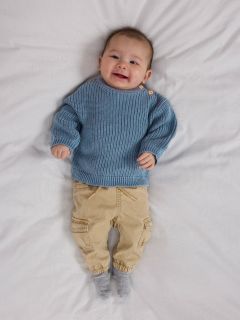 Un bébé souriant est allongé sur une couverture blanche, portant un pull en tricot bleu, un pantalon en velours côtelé marron avec des poches et des chaussettes grises. Le bébé a l'air heureux et détendu, avec les bras légèrement pliés au niveau des coudes.