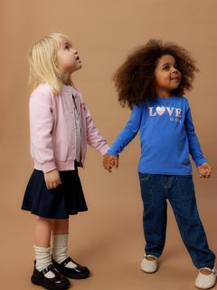 Deux jeunes enfants se tiennent par la main devant un fond marron uni. Un enfant porte une veste rose et une jupe, l'autre un jean bleu et une chemise bleue avec l'inscription "L'AMOUR EST ENFANT". Les deux regardent vers le haut avec curiosité.