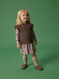Une petite fille aux cheveux blonds se tient souriante devant un fond vert, portant un pull sans manches marron sur une robe à fleurs, des chaussettes montantes et des chaussures marron.