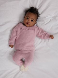 Un bébé est allongé sur une couverture blanche et porte un pull en tricot rose clair avec un col Claudine et un pantalon assorti, ainsi que des chaussettes couleur crème.