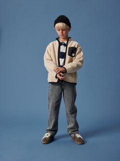 Un garçon aux cheveux blonds porte un bonnet noir, une veste polaire crème sur une chemise rayée, un jean gris et des baskets. Il se tient devant un fond bleu uni et regarde vers le bas, les mains jointes.