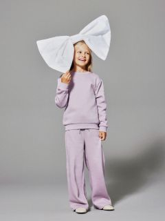 Une jeune fille souriante en survêtement lavande se tient devant un fond gris et porte un très grand nœud blanc sur la tête.