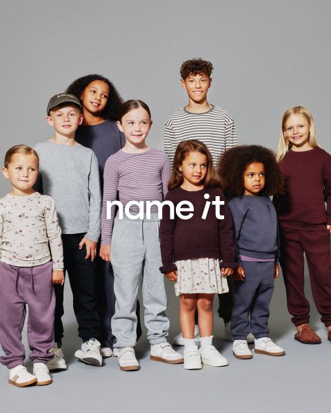 Un groupe de huit enfants différents se tient ensemble devant un fond gris, tous portant des tenues décontractées et assorties dans des tons neutres et pastel. Le texte "name it" apparaît au centre de l'image.