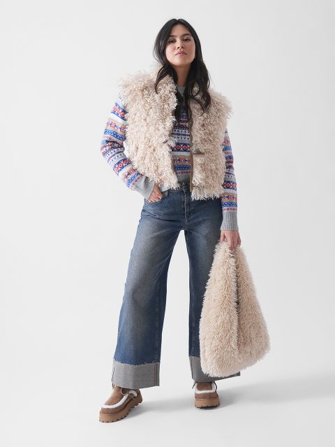Une femme se tient avec assurance devant un fond blanc uni, vêtue d'un gilet beige duveteux, d'un pull à motifs, d'un jean à jambes larges, de chaussures à plateforme marron clair et d'un sac beige duveteux assorti.