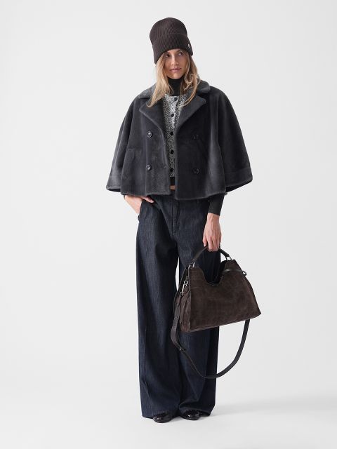Une femme se tient devant un fond blanc, portant un bonnet marron foncé, un manteau de fourrure gris court, un jean foncé à jambes larges et des chaussures noires, et tenant un grand sac à main marron foncé.