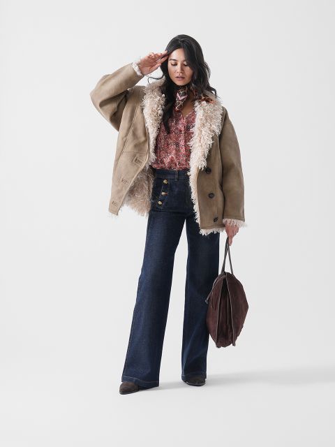 Une femme pose en veste en peau lainée marron clair sur une chemise à motifs, un jean évasé taille haute et des bottes marron, tenant un grand sac marron dans sa main gauche devant un fond blanc uni.