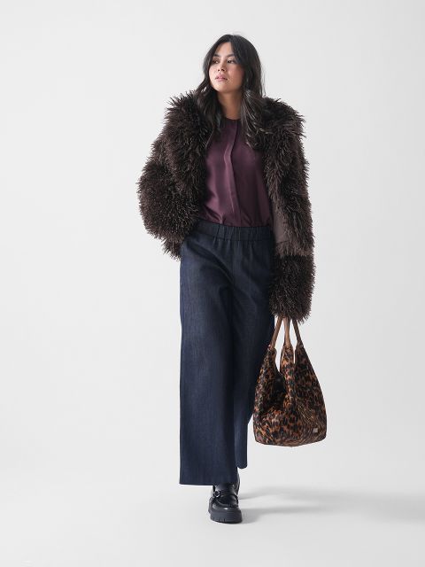 Une femme porte une veste en fausse fourrure marron hirsute, un chemisier violet, un jean foncé à jambes larges et des chaussures noires, tout en portant un sac fourre-tout à motif léopard devant un fond blanc uni.