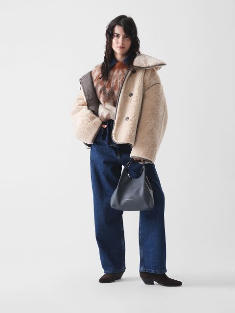 Une femme se tient devant un fond simple, portant une veste en peau lainée beige, un châle en fourrure marron, un jean bleu ample et des bottes à talons hauts foncées, et tenant un sac à main noir dans sa main gauche.