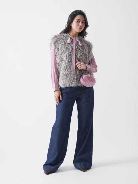 Une femme se tient devant un fond blanc et porte un chemisier rose avec un nœud, un gilet en fausse fourrure grise, un jean bleu ample, des chaussures foncées et un petit sac à main rose.