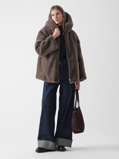 Une femme portant un manteau marron à capuche en fausse fourrure, un jean foncé à jambes larges et à revers, des chaussures noires et un sac à main marron en daim se tient devant un fond blanc uni.
