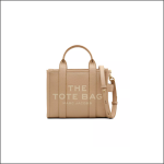 Beigefarbene Marc Jacobs Tote Bag mit zwei Henkeln und abnehmbarem Schulterriemen. Auf der Vorderseite ist der Schriftzug "THE TOTE BAG MARC JACOBS" in Weiß aufgedruckt.