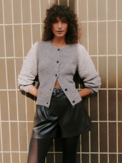 Une femme aux cheveux bouclés se tient devant un mur carrelé. Elle porte un cardigan gris clair duveteux, un short en cuir noir taille haute et des collants noirs, les mains dans les poches et une expression confiante sur le visage.