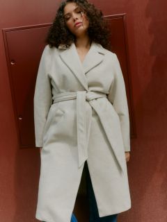 Une personne aux cheveux bouclés et portant une ceinture beige clair se tient devant un mur brun rougeâtre et regarde légèrement vers le bas avec une expression détendue.