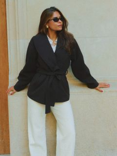 Femme en manteau ceinturé noir, pantalon blanc et lunettes de soleil appuyée contre un mur de pierre beige, une main sur le mur, l'autre sur la hanche, regardant sur le côté.