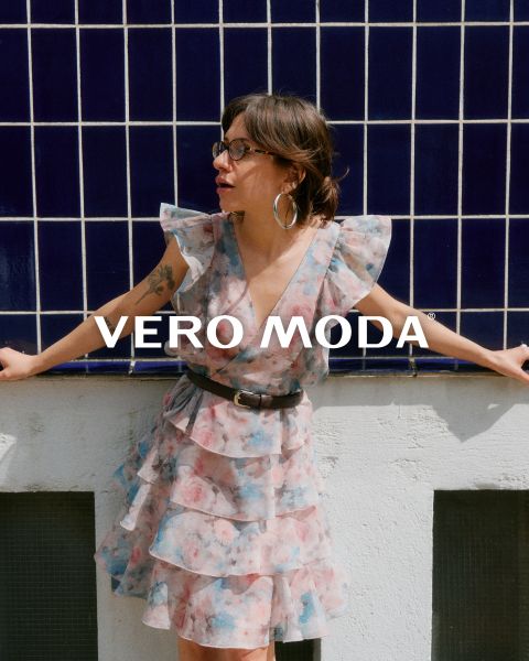 Une femme portant des lunettes et une robe à fleurs à volants est appuyée contre un mur blanc devant un fond carrelé bleu. L'inscription "VERO MODA" apparaît au centre de l'image.