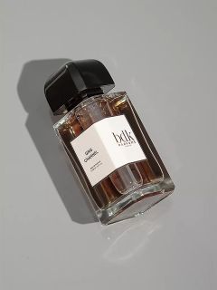 Un flacon de parfum en verre transparent avec un bouchon noir repose sur une surface gris clair. L'étiquette blanche porte les inscriptions "Gris Chanel" et "bdk Parfums". Le flacon projette une ombre douce.