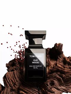 Un flacon noir du parfum Tom Ford Oud Wood est posé sur un morceau de bois texturé, à proximité duquel sont éparpillées de petites graines brunes, le tout sur un fond blanc.