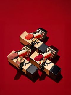 Trois rouges à lèvres dorés et noirs avec différentes nuances de rouge, disposés en diagonale sur un fond rouge uni. L'emballage est orné d'un logo noir sur le boîtier doré.