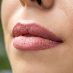 Nahaufnahme der Lippen einer Person, die matten rosa Lippenstift trägt. Die Haut erscheint glatt, mit einem natürlichen Teint und einem Hauch von Lächeln. Der Hintergrund ist sanft verschwommen.
