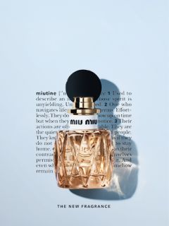 Un flacon de parfum Miu Miu avec un bouchon noir rond est placé devant un texte flou sur fond blanc. Le parfum est de couleur ambre clair, et le texte en dessous indique "LE NOUVEAU PARFUM".