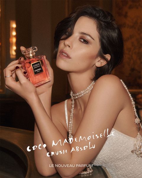 Une femme en robe blanche tient un flacon du parfum « Coco Mademoiselle » ; on peut également voir d'élégants bijoux en perles et un texte publicitaire pour le parfum.